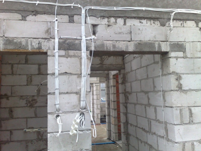 Instalacja elektryczna w surowym wnętrzu budynku z białych bloczków, widoczne kable prowadzone po ścianach i w otworach, przygotowane pod gniazdka i włączniki.